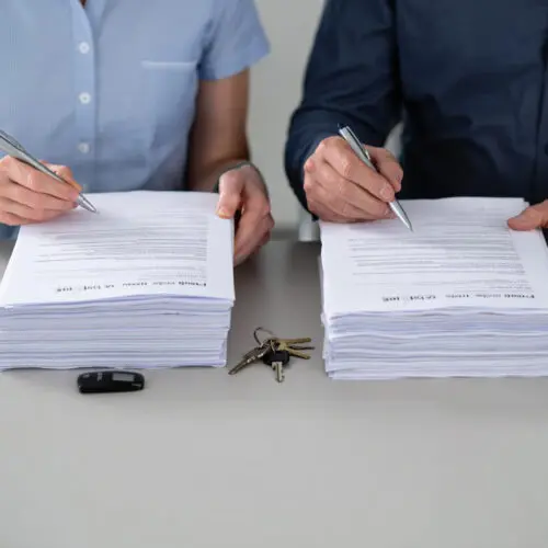 Zwei Personen unterzeichnen gleichzeitig separate, identische Vertragsstapel; ein Schlüsselbund und ein Transponder liegen dazwischen.